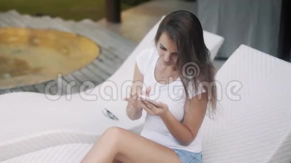 年轻的美女坐在泳池旁的躺椅上用电话享受时光视频的预览图