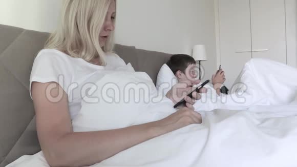 母亲和儿子坐在床上使用智能手机娱乐和社交交流隔离期间的联系视频的预览图