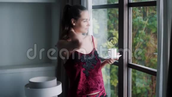 穿着睡衣的年轻女子早上喝着咖啡看着窗外视频的预览图
