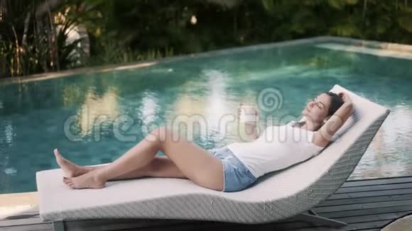 穿着休闲装的年轻女子躺在靠近游泳池的日光浴床上喝咖啡视频的预览图
