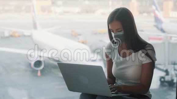 戴防护面罩的女人坐在机场的窗户附近使用笔记本电脑视频的预览图