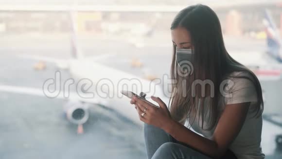 戴防护面罩的女人坐在机场的窗户附近使用智能手机视频的预览图