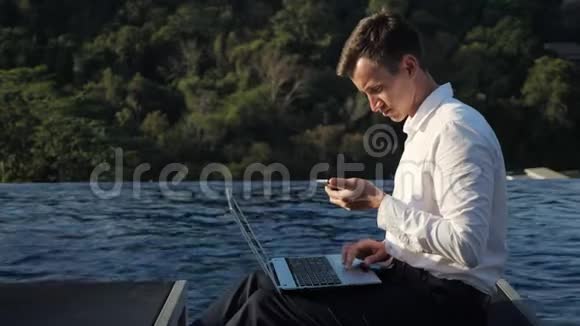 男人在笔记本电脑上工作在森林和游泳池旁看电话视频的预览图