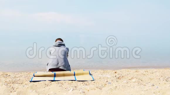 一个孤独悲伤的女孩穿着灰色的帽衫坐在海滩上背面视频的预览图