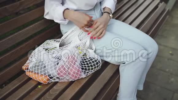年轻女子坐在长凳上在夏天的公园里从棉网购物袋里拿番茄零浪费购物视频的预览图