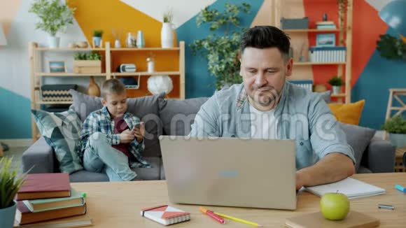 中年男子在家里使用笔记本电脑而儿子则忙于智能手机视频的预览图