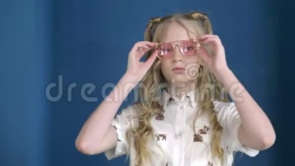 可爱的少年女孩卷曲的金发透过粉红色太阳镜在蓝色工作室穿着粉色衣服的时尚女孩视频的预览图