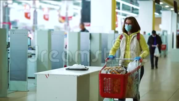 戴着面具的女人带着装满食物的购物车沿着收银机走视频的预览图