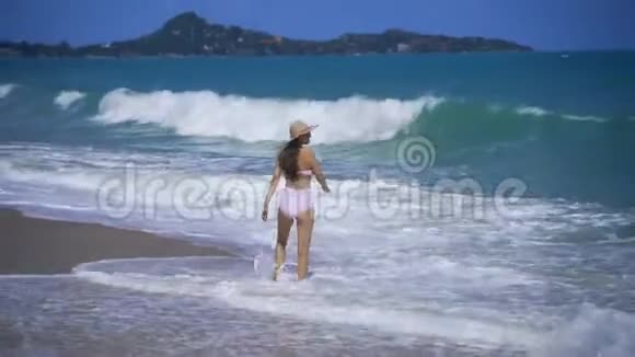 一个穿着粉红色泳衣的漂亮女孩沿着沙滩散步巨大的海浪在海滩上翻滚视频的预览图