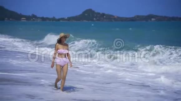 一个女孩沿着海洋走看着海浪一个戴太阳镜和粉红色泳衣的女孩视频的预览图