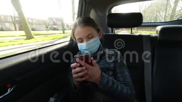 戴口罩的无聊病女孩坐在汽车后座看着智能手机视频的预览图
