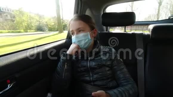 戴口罩的无聊病女孩坐在汽车后座看着智能手机视频的预览图