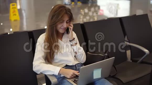 穿着白色衬衫的漂亮女孩在候机室等候登机时会使用手机和笔记本电脑在机场工作视频的预览图