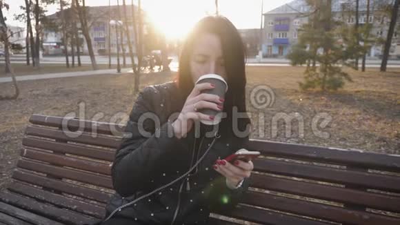 漂亮的女人用电话坐在有咖啡杯的公园长凳上视频的预览图