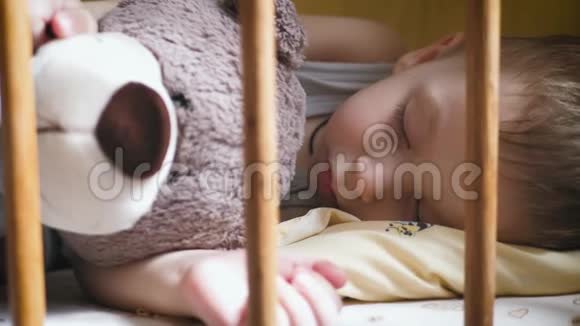 小可爱的蹒跚学步的男孩和泰迪熊睡在儿童床上视频的预览图
