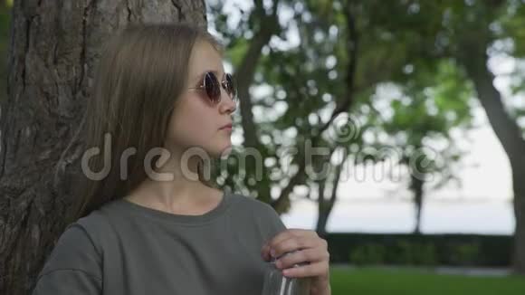 太阳眼镜的年轻女孩在夏天公园户外喝橙汁视频的预览图