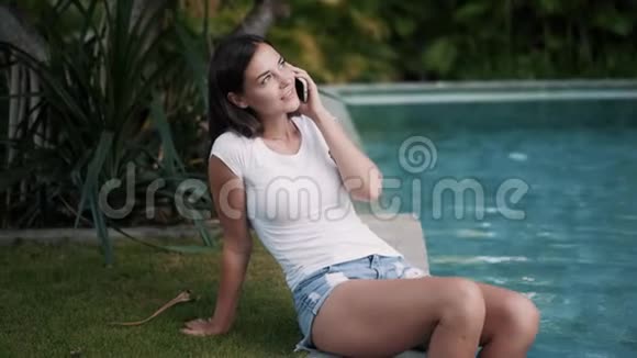 年轻女子坐在池边双脚在水中打电话视频的预览图