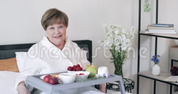 微笑的美丽老太太在床上吃健康早餐的画像穿着长袍的快乐成熟女士躺在床上视频的预览图