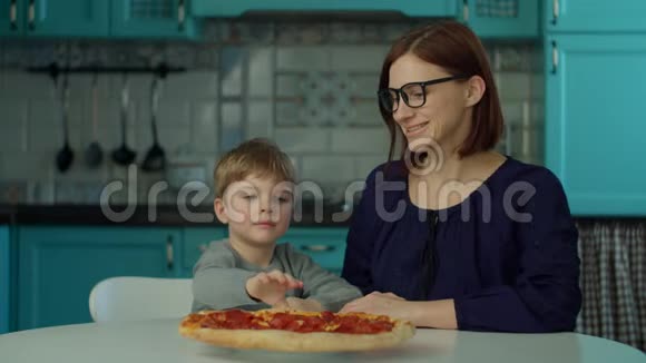 幸福的母子一家在蓝色厨房的家里吃披萨30个戴眼镜的女人和金发男孩一起享受披萨视频的预览图
