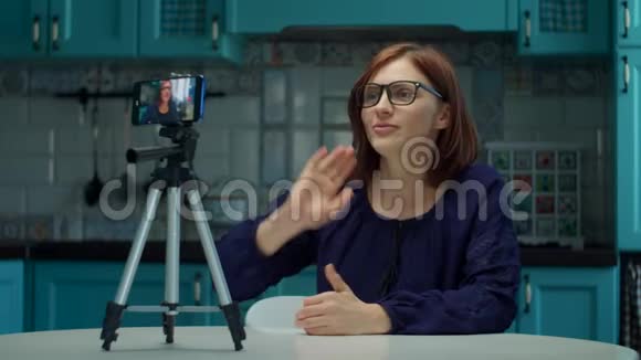 年轻的30岁的女性博主在蓝色厨房的家里用手机录制视频在家工作的戴眼镜的女人视频的预览图