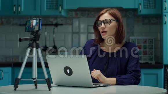 年轻的30岁的女性博主在蓝色厨房的家里用手机录制视频在家工作的戴眼镜的女人视频的预览图
