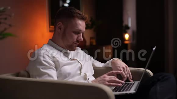 坐在舒适的客厅里的一个自信的留着胡子的年轻人在家里坐在软扶手椅上用笔记本电脑视频的预览图