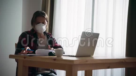 戴着医用口罩在家工作的年轻女人用她的笔记本电脑视频的预览图