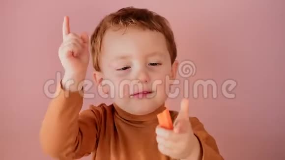可爱的小红发男孩在粉红色的背景上吃胡萝卜视频的预览图