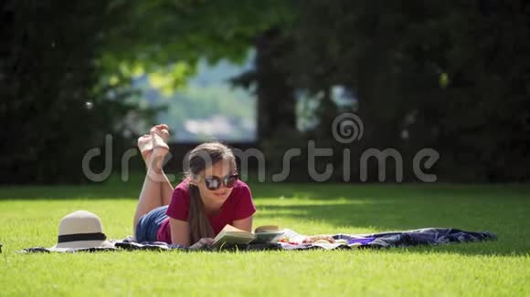 阳光明媚的一天在花园里看太阳眼镜书的漂亮女孩野餐休息晒太阳视频的预览图