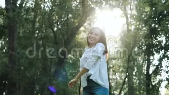 一个穿着白色衬衫的迷人女孩在公园的树木之间的街道上盘旋的前景快乐视频的预览图