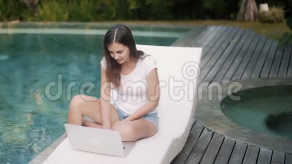 女性自由职业者坐在游泳池附近的日光浴床上在笔记本电脑上工作收到好消息视频的预览图