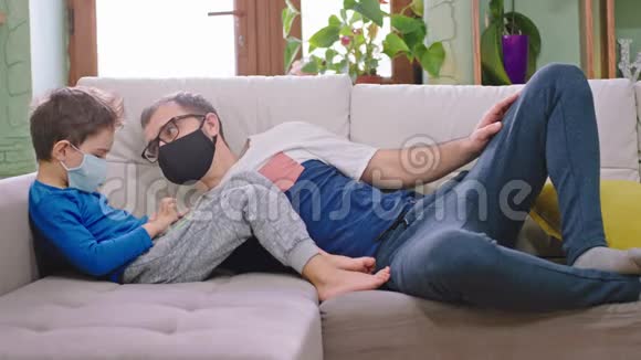 在家里的沙发上爸爸和他的小儿子在一起戴着防护面具他们用智能手机来做防护视频的预览图