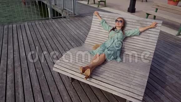 穿着蓝色衣服的女人太阳眼镜放松双臂伸展在木制太阳躺椅上视频的预览图