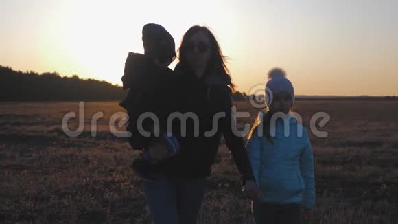 快乐的年轻母亲和两个孩子在日落时漫步在草地上友好家庭概念的剪影视频的预览图