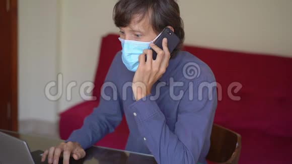 一个戴着面罩的年轻人在冠状病毒自我隔离期间在家工作他用手机说话慢动作视频的预览图