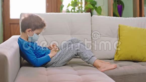 带着防护面具的小男孩带着智能手机坐在沙发上玩着隔离概念的游戏视频的预览图