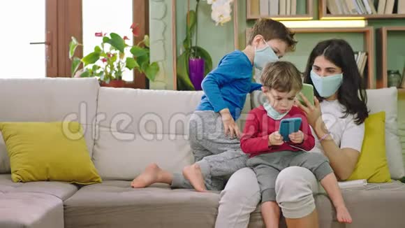 母亲带着两个带防护面罩的孩子坐在沙发上他们呆在家里用隔离器隔离视频的预览图