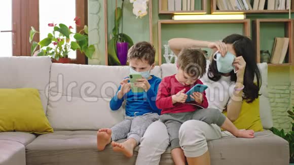 年轻的妈妈带着她的两个儿子在沙发上戴着防护面具而正在隔离孩子们玩智能手机视频的预览图