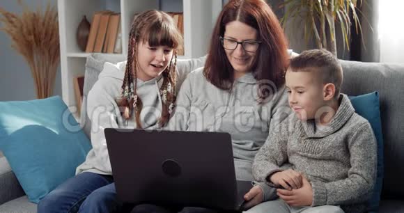微笑的母亲和孩子们用笔记本电脑在沙发上放松视频的预览图