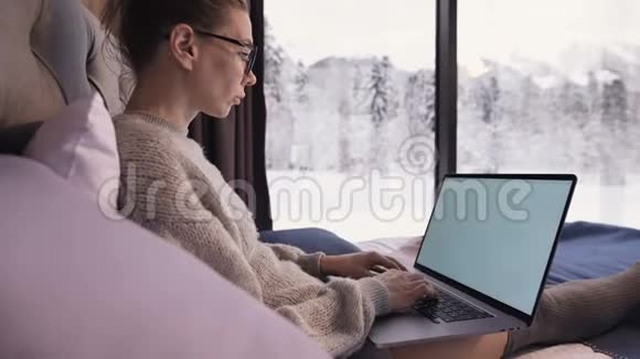一个戴着眼镜的女自由职业者坐在乡间别墅的床上房子后面有一扇雪亮的窗户视频的预览图