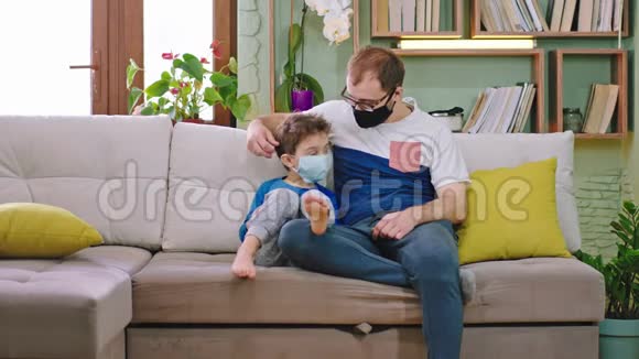 冠状病毒爸爸和他的小儿子在沙发上呆在家里隔离他们戴着防护面具给了一个视频的预览图