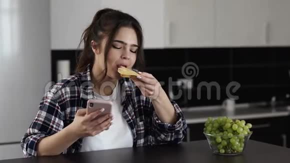 检疫期间厨房里的年轻女子坐在桌子旁吃奶酪三明治在智能手机上聊天社会视频的预览图