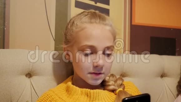 可爱的微笑少女与金发辫子使用移动沙发一个漂亮女孩在看智能手机屏幕视频的预览图