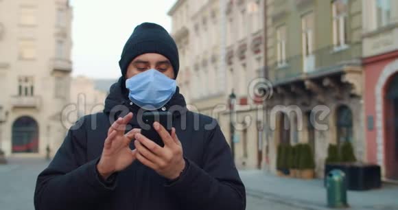一个人站着的时候戴着医用口罩的年轻人用智能手机戴着防护面罩的人看着视频的预览图