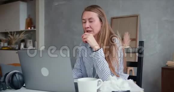 美丽的年轻白种人金发女人患感冒打喷嚏同时使用笔记本电脑在家工作隔离视频的预览图