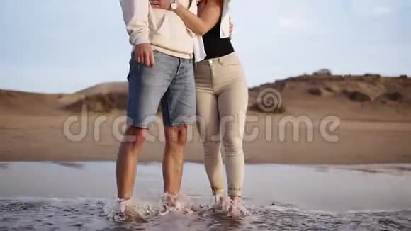时髦的年轻夫妇站在沙滩海岸线上观看来自海洋的日落波浪穿着太阳镜和休闲装视频的预览图