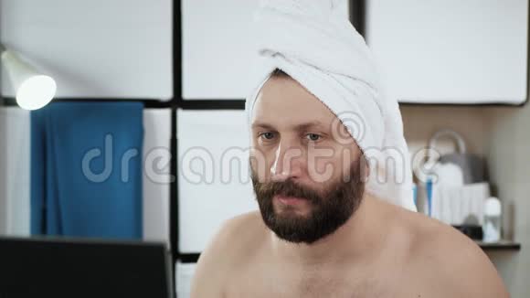 男人把补丁贴在脸上一个穿毛巾戴头巾的可爱男人看着镜子和化妆品视频的预览图