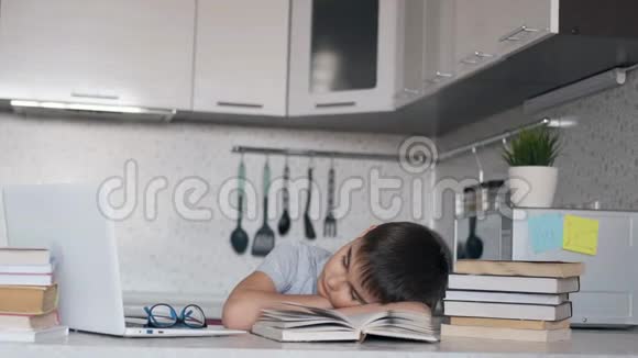 疲惫的小学生坐在笔记本电脑和教科书旁边的桌子上做作业睡着了左摄像机移动视频的预览图