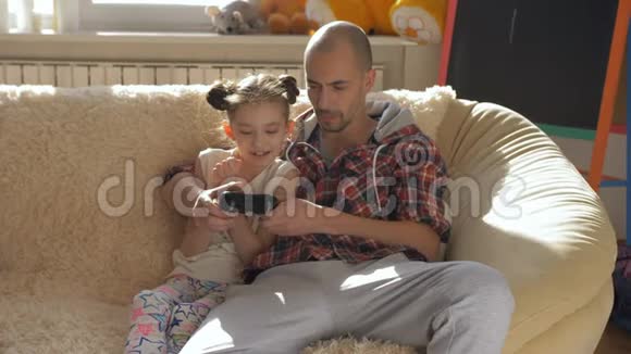 爸爸和女儿一起玩电子游戏坐在阳光房的沙发上视频的预览图