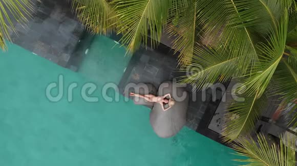 穿着米色比基尼和草帽的苗条年轻女子在豪华游泳池放松的无人机空中景观假期概念视频的预览图
